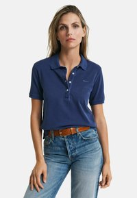 Donkerblauwe polo gemaakt van katoen, met een drieknopenlijst, korte mouwen en een subtiele logo op de linker borst. Gecombineerd met lichte spijkerbroek.