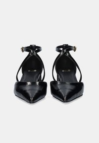Zapatos de tacón alto de charol negro con punta afilada, que cuentan con correas en los tobillos entrecruzadas y un pequeño acento de hebilla dorada.