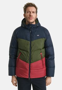 Padded Jacke mit Kapuze, mehrfarbig in Marineblau, Grün und Rot, mit Reißverschlusstaschen und einem Zickzackmuster über der Brust.