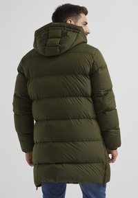 Grüne Daunenjacke mit Kapuze, gefüttert, längerer Rücken und seitliche Reißverschlüsse. Glattes, wasserabweisendes Material.