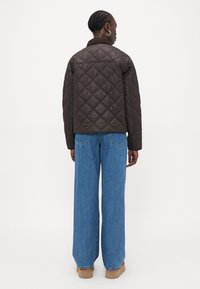 Veste marron matelassée avec un motif en losanges, dotée d'un col haut et d'une coupe amples, associée à un jean bleu clair à jambes larges et à des chaussures beiges.