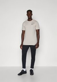Camiseta de color beige claro con un pequeño logo negro de Nike, combinada con pantalones ajustados negros y zapatos deportivos negros. Calcetines blancos visibles.