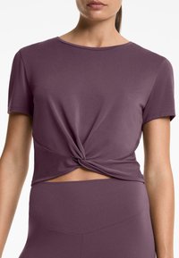 Fialový crop top z měkké tkaniny, s předním uzlem a výřezem v pase. Krátké rukávy a kulatý výstřih.