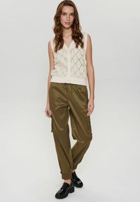 Krämfärgad stickad väst med diamantmönster och knäppning framtill, kombinerad med olivgröna cargobyxor och svarta platåskor.