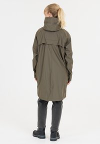 Lang olivengrøn regnfrakke med høj krave og bagventil, parret med mørkegrå bukser og sorte støvler. Glat, vandtæt materiale.