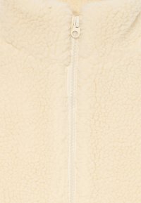 Krémová fleecová bunda s centrálním zipem. Materiál má texturovaný, nadýchaný vzhled a jemný povrch.