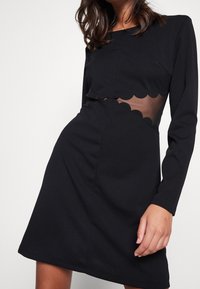 Abito nero con maniche lunghe, corpetto aderente, tagli ondulati in vita e una texture liscia.