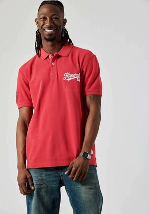 Polo shirt rouge avec un col, arborant une broderie de logo blanche sur la poitrine gauche. Porté avec un jean en denim bleu.
