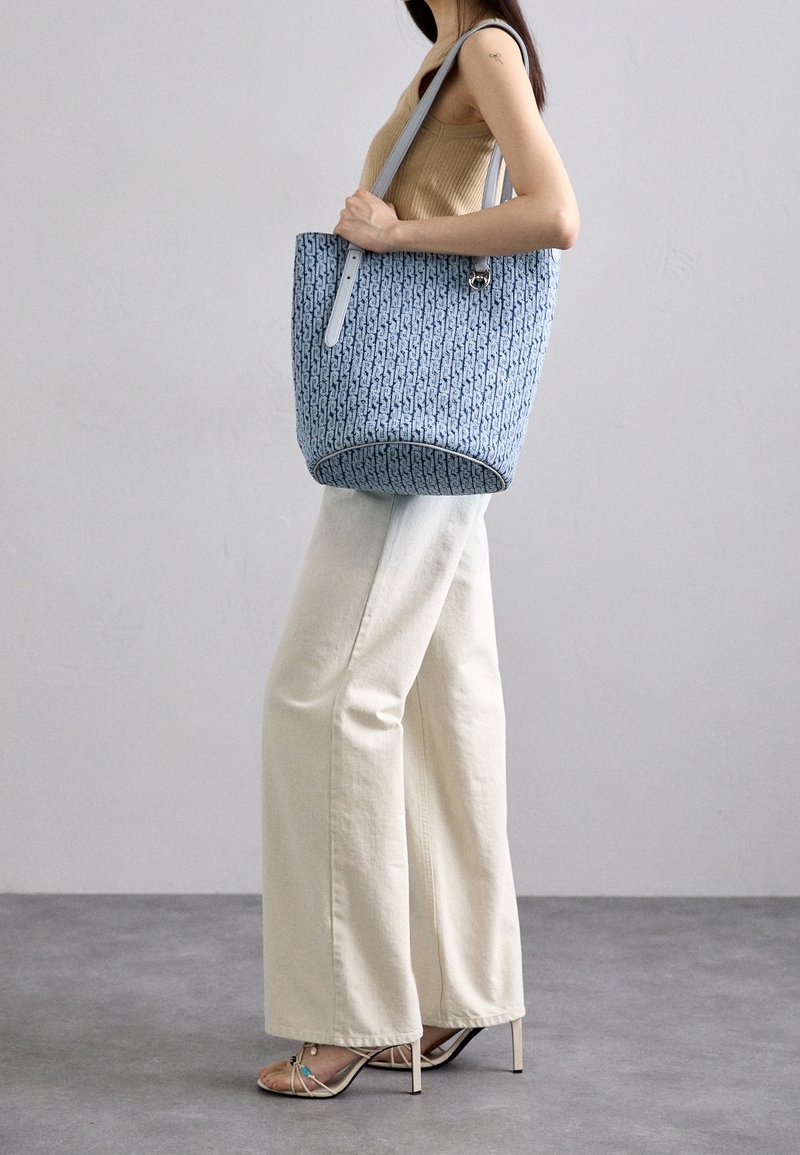 Blue patterned tote bag with leather straps, featuring a rounded base and open top, worn with light-colored wide-leg pants.