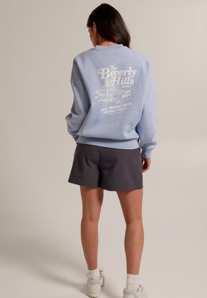 Vrouw staat met de rug naar de camera, draagt een lichtblauwe sweatshirt met de tekst "Beverly Hills Resort" en donkergelige korte broek, voor een effen achtergrond.