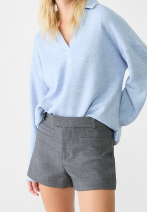 Mujer con suéter azul claro de manga larga metido en pantalones cortos grises a la cintura hechos a medida, sobre un fondo liso.