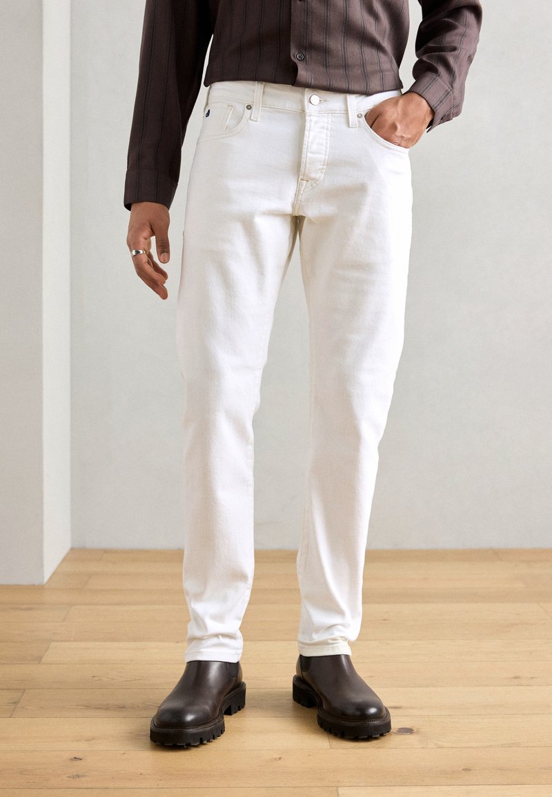 Homme portant un jean blanc slim, des bottes noires robustes et une chemise rayée marron foncé, avec une main dans la poche sur un sol en bois.