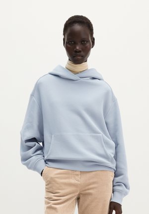 Femme portant un sweat à capuche bleu clair sur un col roulé beige et un pantalon beige en velours côtelé, debout avec une main dans la poche sur un fond uni.
