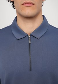 Blå polo-skjorte laget av glatt stoff, med en kort glidelås ved halsen og et klassisk krage-design. Ingen mønstre eller aksenter synlige.