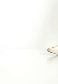 Osoba obuvající bílé tenisky, která kráčí vpřed po hladkém bílém povrchu na pozadí jednobarevné bílé barvy.