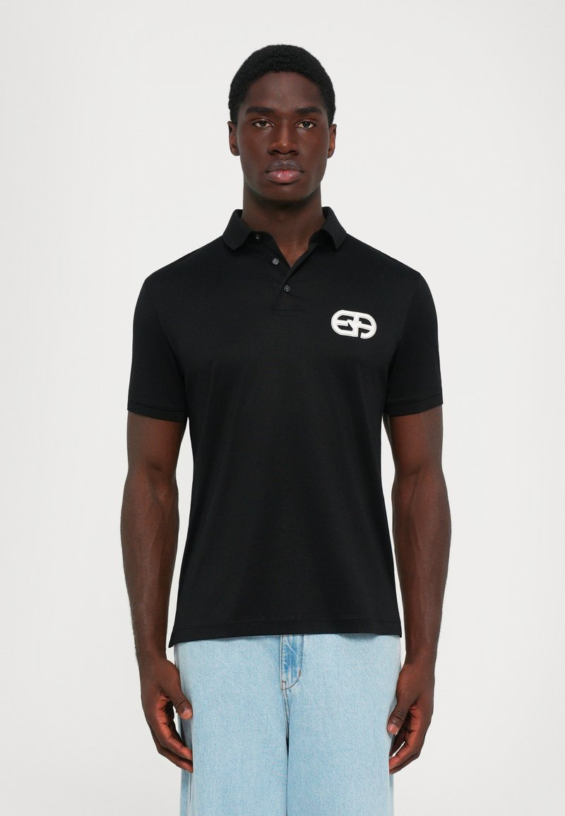 Bărbat îmbrăcat cu un tricou polo negru cu mâneci scurte, având un logo alb pe pieptul stâng și blugi de culoare albastru deschis, stând în fața unui fundal alb.