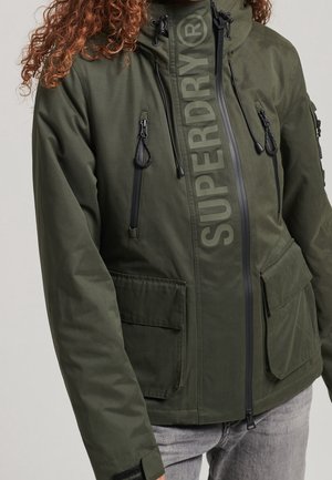 Persona vistiendo una chaqueta verde oliva de Superdry con múltiples bolsillos con cremallera y solapa, de pie frente a un fondo liso.