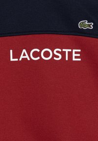 Felpa rossa e blu navy con la scritta "LACOSTE" in bianco. Presenta un piccolo logo del coccodrillo verde sulla parte superiore destra. Tessuto liscio.