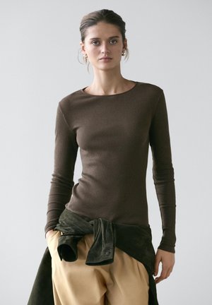 Femme à la peau claire et aux cheveux foncés portant un haut marron ajusté à manches longues, un pantalon beige et une veste foncée nouée autour de la taille.