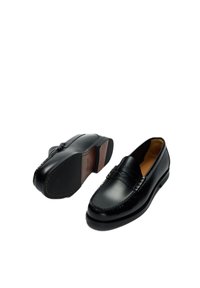 Ζευγάρι μαύρα δερμάτινα παπούτσια τύπου penny loafer, το ένα όρθιο και το άλλο που δείχνει την υφή της σόλας, πάνω σε λευκό φόντο.