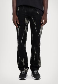 Melnas džinsi ar saistītu krāsu efektu, kas satur baltas svītras. Taisna kāju dizains ar redzamām šuvēm, apvienoti ar melnām sporta apaviem.