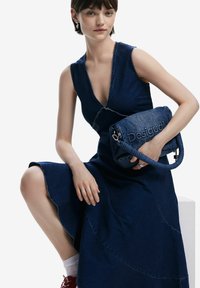 Jeanshandväska med flätat handtag, prydd med "Desigual"-logotypen. Modellen bär en ärmlös blå denimklänning med en utsvängd nederdel.