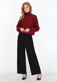 Bordeauxfarbener Rollkragenpullover mit puffigen Ärmeln, kombiniert mit hoch taillierten schwarzen weiter geschnittenen Hosen und schwarzen hohen Schuhen. Glattes Material.