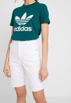 Kvinde iført teal Adidas t-shirt og hvidt denimshorts med høj talje og frynset kant, stående mod en ensartet lys baggrund.