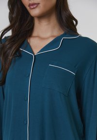 Camicia azzurro-verde con taschino, piping bianco lungo il colletto e il taschino, texture a costine e vestibilità rilassata.