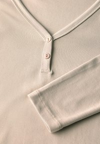 Haut beige clair à manches longues avec un décolletage en V, deux boutons pâles à l'avant et une texture côtelée douce. Le tissu semble extensible et lisse.