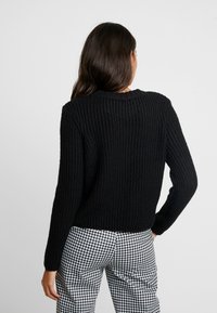 Schwarzer gestrickter Pullover mit gerippter Textur, rundem Ausschnitt und langen Ärmeln, kombiniert mit gemusterten schwarz-weißen karierten Hosen.