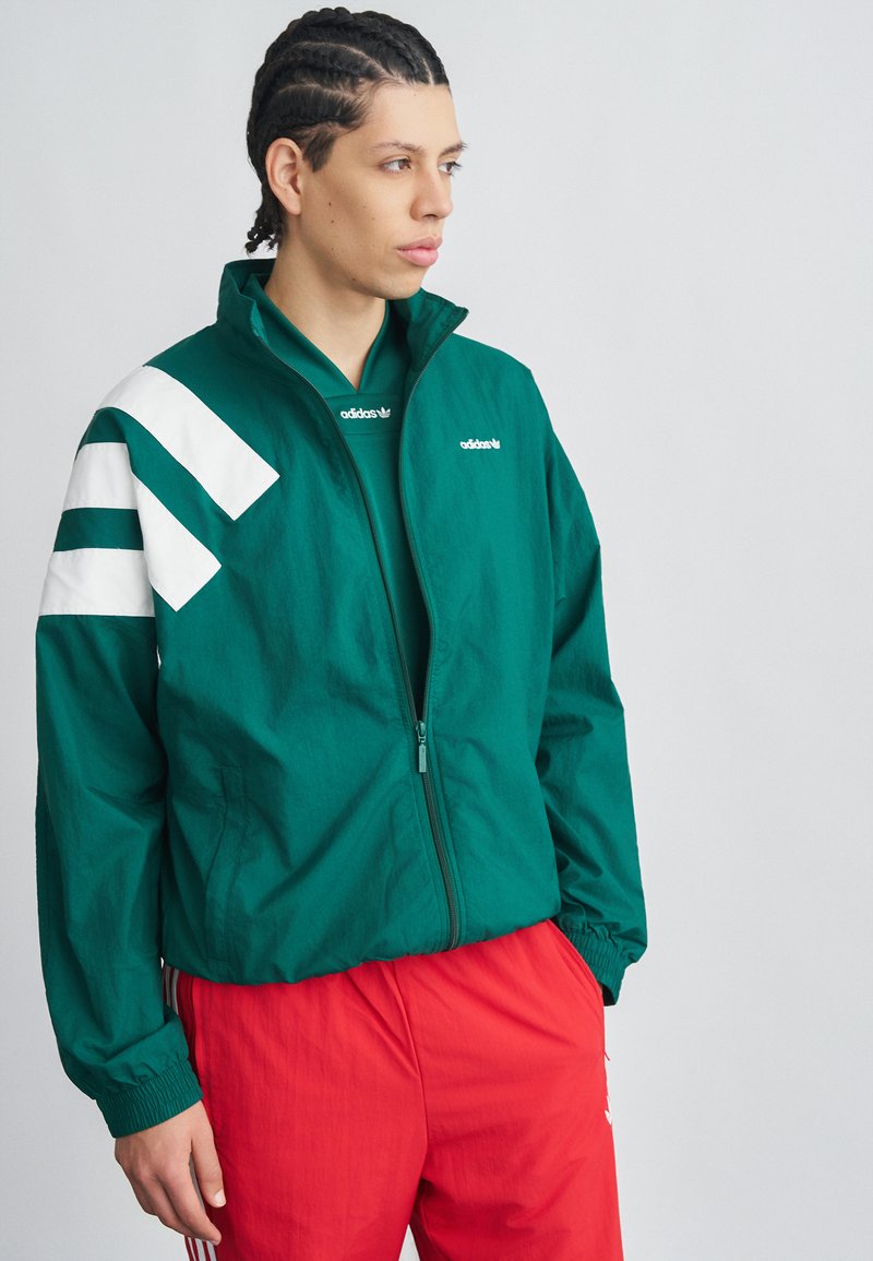 Mladá osoba s vrkočmi, oblečená v zelenej bunde Adidas s bielymi pruhmi a červených nohaviciach Adidas, pozerajúca sa do strany na jednoliatom pozadí.