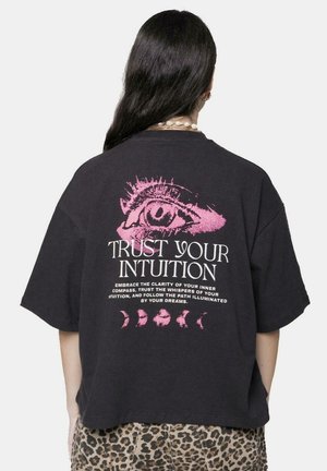 Camiseta negra de gran tamaño con un destacado gráfico de un ojo rosa y el texto "CONFÍA EN TU INTUICIÓN" con fases lunares decorativas debajo.