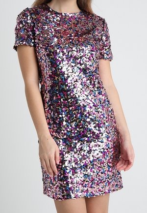 Frau trägt ein knielanges, kurzärmeliges Kleid, bedeckt mit bunten, glänzenden Pailletten, steht mit locker an den Seiten hängenden Armen.