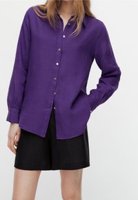 Femme portant une chemise violette à manches longues boutonnée et un short noir, avec une main dans la poche, sur un fond blanc uni.