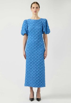 Kvinde iført en tekstureret, lys blå midikjole med puffede korte ærmer og sorte spidse hæle, stående mod en ensartet lys baggrund.