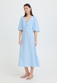 Femme portant une robe midi bleu clair avec des manches volantes, un décolleté en V plissé, et des sandales à talons argentées, debout sur un fond blanc.