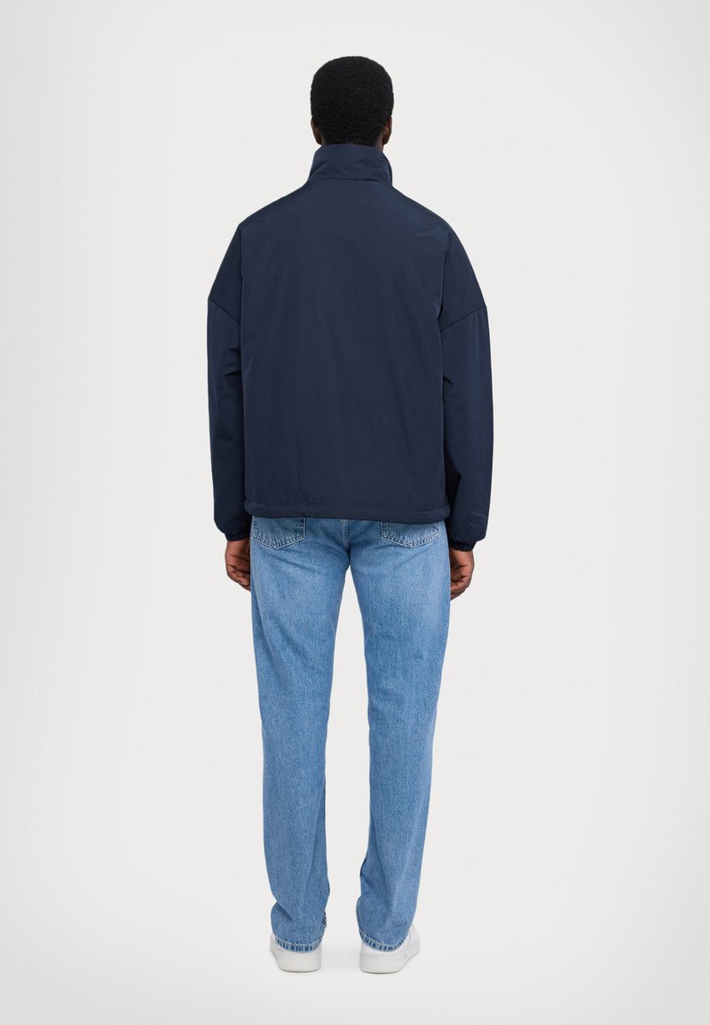 Chaqueta navy con cuello alto, corte oversize y textura suave, combinada con jeans de color azul claro y zapatillas blancas.
