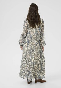 Lang, svævende kjole i let stof med et creme- og marineblåt paisley-mønster. Den har lange ærmer og en lagdelt nederdel. Bæres med brune ankelstøvler.