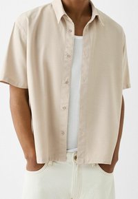 Koszula z krótkim rękawem na guziki w jasnobeżowym materiale o miękkiej teksturze, wyposażona w klasyczny kołnierzyk i subtelne detale z guziczków.