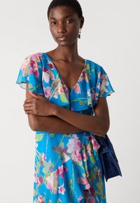 Abito floreale blu con maniche corte a volant, caratterizzato da motivi di fiori rosa e verdi. Abbinato a una borsa a mano blu scuro.