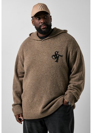 Mand iført en brun hættetrøje med sort logo og beige baseballkasket, stående med den ene hånd i lommen og kigger til siden.