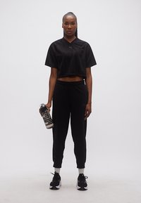 Αθλητικό άτομο που στέκεται κρατώντας ένα μπουκάλι νερό, φορώντας μαύρο κοντό τοπ, αθλητικές φόρμες, λευκές κάλτσες και μαύρα αθλητικά παπούτσια σε απλό φόντο.