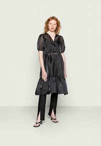 Vestido de cetim preto com mangas bufantes e bainha com babados, combinado com calças de cetim preto com fendas laterais e sandálias de salto agulha.