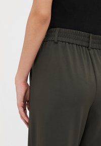 Tumedat rohelised, elastse vöökohtaga püksid sileda tekstuuriga, tagaküljel dart'idega ja küljetaskutega, nähtavad tagantpoolt.