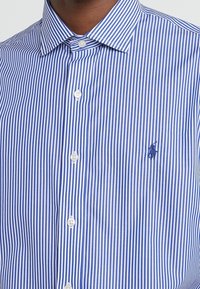 Camicia a maniche corte a righe con strisce verticali blu e bianche, colletto con bottoni e piccolo logo ricamato sul petto.