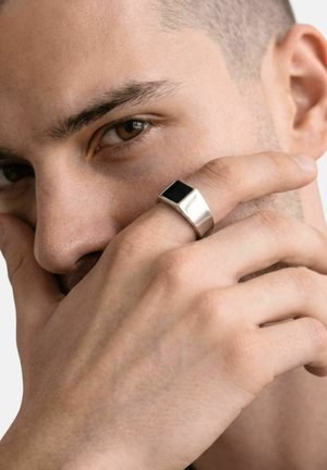 Close-up van een persoon die deels zijn gezicht bedekt met een hand met een brede zilveren ring met een zwarte vierkante steen.