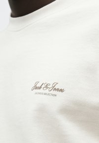 Ανοιχτό μπεζ μπλουζάκι από μαλακό βαμβάκι, με ριμπ λεπτομέρεια στο λαιμό και καφέ κεντημένο λογότυπο της μάρκας στην αριστερή πλευρά του στήθους.