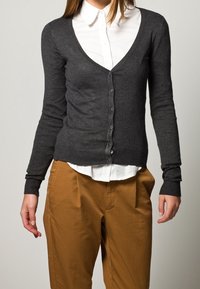 Grauer Strickcardigan mit tiefem V-Ausschnitt, Knopfverschluss und gerippten Bündchen. Kombiniert mit einem weißen Hemd mit Kragen und braunen Hosen.
