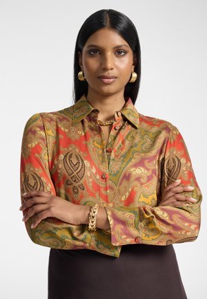 Femme aux longs cheveux noirs, debout les bras croisés, portant une chemise rouge et or à motifs cachemire, des boucles d'oreilles anneaux dorés, un collier chaîne et un bracelet.